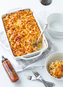 A baked macaroni and cheese casserole sits in a white dish with a serving spoon, alongside a bowl of macaroni, empty bowls, a bottle of Terrapin Ridge Farms Truffle Hot Sauce, red wine, and forks on a white surface.