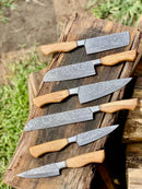 Five Texan Knives Sharp Kitchen Knife Sets, each with a 13" Damascus steel blade, olivewood handle, and leather sheath, are arranged outdoors on rustic wood with grass and timber in the background.