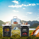The DirectDeals Folding Camping Table with Storage Organizer is set up outdoors on grass, holding cookware, dishes, and fresh produce. Nearby are a camping stove, an orange tent, mountains, and a lake under the blue sky.
