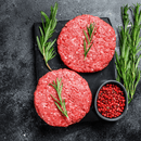 Two raw American ground beef burger patties from the Dude Food Gentleman’s Meat Box by Dude Food rest on a black cutting board, each topped with a fresh sprig of rosemary. Fresh rosemary and a small bowl of pink peppercorns are arranged beside the patties on a dark surface.