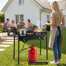 A woman cooks on the DirectDeals 2 Burner Propane Stove 160000 BTU with Adjustable Regulator in a backyard outdoor kitchen as four friends chat nearby, with a red gas cylinder below and a white house in the background.