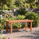 The DirectDeals 2-Person Outdoor Wood Bench with Slatted Seat - Set of 2 rests on a gravel path in a garden filled with flowers, bushes, and trees, with a stone wall in the background.