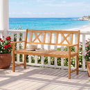 A 2-Person Acacia Wood Garden Bench with X-Shaped Backrest by DirectDeals sits on a porch among potted red and pink flowers, overlooking the beach under a clear blue sky.