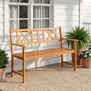 A DirectDeals 2-Person Acacia Wood Garden Bench with X-Shaped Backrest sits on a woven rug outside a white house, surrounded by potted ferns and blooming flowers near two windows.