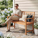A man in sunglasses, holding a mug, sits on the DirectDeals 2-Person Acacia Wood Garden Bench with X-Shaped Backrest, surrounded by hydrangeas and potted plants on a stone patio beside a beige house.