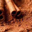 A close-up of two cinnamon sticks resting on a pile of The Spice Lab Organic Ground Vietnamese Saigon Cinnamon, highlighting the warm brown color and fine texture of both the sticks and the organic ground cinnamon powder.