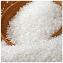 A close-up of a wooden spoon filled with The Spice Lab Sanding Con AA Sugar—coarse white crystals—with more sugar scattered underneath, perfect for decorative baking.
