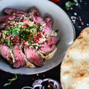 Sliced rare steak on a plate with creamy sauce, garnished with pomegranate seeds and herbs, served alongside a piece of flatbread. The dish is subtly enhanced with The Spice Lab Organic Ground Coriander, imparting warm, aromatic notes reminiscent of traditional Indian cooking.