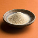 A brown ceramic bowl filled with a mound of pale yellow The Spice Lab Organic Granulated Onion, a classic pantry staple, is placed on an orange surface.