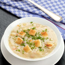 A bowl of creamy soup topped with croutons and a sprinkle of The Spice Lab Fancy Chervil sits on a white plate, with a blue and white checkered napkin and a spoon in the background.