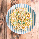A plate of creamy pasta with zucchini slices is finished with a sprinkle of The Spice Lab Green Chives by The Spice Lab, and served on a blue and white striped plate atop a rustic wooden table.