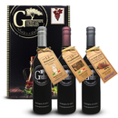 Three bottles from the Georgetown Olive Oil Co. 3 Bottle Aged Balsamic Vinegar Gift Set are artfully displayed with brown and beige tags in front of a black gift box adorned with the Georgetown Olive Oil Co. logo and a grape illustration. Each tag provides details about the type and age of the aged balsamic vinegar within, making this set an exceptional gourmet gift for any occasion.