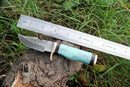 The Texan Knives Custom Handmade Best Survival Knife by Texan Knives features a patterned Damascus steel blade and turquoise handle. Shown on a tree stump with a metal ruler above it, this survival knife measures about 6 inches long.