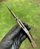 A black-gloved hand holds the Texan Knives Custom Handmade Damascus Blade EDC Folding Pocket Knife, featuring a color bone handle and engraved brass bolster, partially open against a blurred green outdoor background.