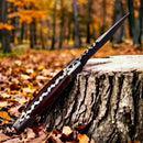 The Texan Knives Small Bushcraft Knife features a 9" Damascus steel blade and rosewood handle, shown upright in a tree stump amid autumn leaves, with its leather sheath nearby.