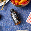 A jar of Spice Witch Chili Oil by Spice Witch sits on a blue textured surface beside a bowl of cherry tomatoes and mini bell peppers, with a red gingham napkin and plate in the background, suggesting its rich savory flavor.