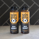 The Spice Witch Hot Honey Duo features two bottles with black caps and orange labels showing a stylized eye, displayed side by side on a gray surface against a diagonal black-tiled background—ideal for sweet and spicy chili honey lovers.