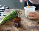 A person stirs ingredients in a plastic container on a wooden cutting board beside a box of 2 Spice Witch Chili Crisp jars and fresh green onions.
