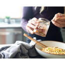 A person scoops Spice Witch Chili Crisp from a Spice Witch Chili Crisp Box of 2 jar onto noodles, with chopsticks on the bowl and a kitchen towel nearby.