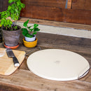 The WPPO LLC Direct 15" Premium Pizza Baking Stone with Handles rests on a rustic wooden counter next to a cutting board with a knife, flour dusted across the surface, and two small potted plants—perfect for recreating delicious brick oven-style pizza at home.