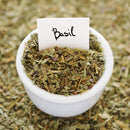 A white bowl filled with The Spice Lab Organic Sweet Basil sits on a surface sprinkled with more dried basil from The Spice Lab. Perfect for Italian-inspired cooking, a small white card labeled “Basil” is placed in the bowl.