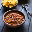A wooden bowl brimming with chili made of beans and ground meat sits on a table beside tortilla chips and two wooden spoons, its hearty aroma elevated by a sprinkle of The Spice Lab Scotch Bonnet Sea Salt for an irresistible Caribbean-inspired kick.
