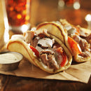 Two gyros filled with sliced meat, tomatoes, and creamy white sauce—each seasoned with The Spice Lab Mediterranean Citrus Seasoning—sit on brown paper, bursting with bold Greek-inspired flavor from The Spice Lab. A small metal cup of extra sauce is on the side for dipping, while a blurred drink can be seen in the background.