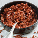 A metal spoon scoops medium grain crystals of The Spice Lab Hawaiian Red Alaea Sea Salt from a dark ceramic bowl set on a white wooden surface, showcasing the salt’s earthy flavor.