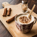 A patterned mug of frothy coffee topped with cinnamon and a hint of The Spice Lab Organic Ground Nutmeg sits on a wooden board, beside three cinnamon sticks and a cinnamon shaker.
