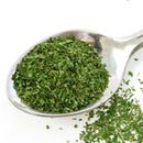 A metal spoon filled with The Spice Lab Organic Parsley Flakes—ideal for garnishing—rests on a white surface, with additional flakes from The Spice Lab scattered beside it.