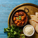 A bowl of chicken curry infused with The Spice Lab Organic Ground Cumin and garnished with cilantro, served with white rice, naan bread, and fresh cilantro on a brass tray over a blue wooden table.