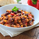 A bowl of chili with ground meat, kidney beans, corn, and diced red peppers is subtly enhanced with a sprinkle of The Spice Lab Ground Oregano—a classic Mediterranean herb from The Spice Lab—and garnished with a sprig of parsley. It sits on a white napkin on a wooden table.