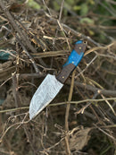 The Texan Knives Gutting Knife—10" handmade Damascus steel hunting knife with a rosewood or resin handle and leather sheath—rests outdoors on dry branches, highlighting its unique patterned blade.