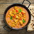 A black bowl of chicken curry, richly seasoned with The Spice Lab Organic Indian Curry Powder and garnished with parsley and red chili slices, served on a wooden table with two pieces of flatbread on the side.