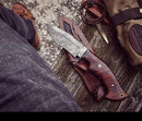 A person in jeans and brown shoes sits outdoors with a Texan Knives Good Hunting Knife—10" Damascus steel blade, rosewood handle, leather sheath—resting on their leg, beside outdoor gear and rope on a wooden surface.