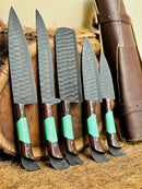 Five Texan Knives’ Premium Knofe Set kitchen knives, featuring 13" forged stainless steel blades and wenge wood with resin handles, are lined up on straw. A brown leather sheath is positioned behind them for stylish storage and display.