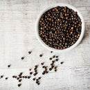 A white bowl filled with The Spice Lab Premium Black Pepper from the Himalayan Pink Salt & Premium Black Pepper Grinder Set sits on a white wooden surface, with some peppercorns scattered outside the bowl.