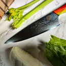 A close-up of the Spaceman Knives EM Wave Chef Knife by Spaceman Knives on a marble surface with celery and lettuce. The blade features a detailed wavy Damascus pattern and a multicolored handle—ideal for any professional kitchen.