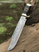 The Texan Knives Authentic Crocodile Dundee Knife features a 16” handmade Damascus steel blade and a buffalo horn or leather handle with a brass guard, displayed outdoors against weathered wood. Includes a leather sheath.