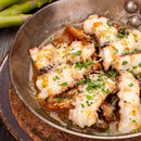A metal pan filled with cooked octopus tentacles in a savory sauce, garnished with chopped parsley and The Spice Lab Whole Leaf Cilantro. Some green asparagus spears are visible in the background.