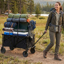 A smiling woman in hiking gear pulls the DirectDeals 38 Inch 400 LBS Heavy-Duty Collapsible Wagon Cart with Adjustable Handle for Camping Garden-Black, loaded with camping gear, across a grassy campsite near a lake and trees.