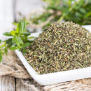 A close-up of The Spice Lab Savory Whole Leaf 1 lb, a square white dish filled with dried, chopped green herbs from The Spice Lab—ideal for foodservice or BBQ seasoning—placed on burlap fabric with fresh herb sprigs in the background.