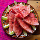 A plate of watermelon slices sprinkled with The Spice Lab Organic Cayenne Pepper, garnished with lime wedges, sits on a wooden table atop a pink woven cloth—a refreshing twist inspired by both Indian and Mexican cooking traditions.