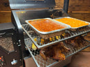 Two metal slide-out racks from the Pimp My Grill® Gen2 Slide-Out Rack System for Green Mountain Grills Ledge / Daniel Boone hold aluminum pans of baked beans and side dishes, with roasted chickens and corn arranged on the lower level.