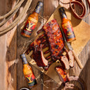 Barbecue ribs on a wooden board, partially sliced, surrounded by two bottles of Elijah's Xtreme Ranchers Revenge (Ghost Pepper) hot sauce from Elijah's Xtreme. Ropes, horseshoes, and a knife are also arranged on the rustic wooden table.