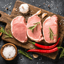 Three raw pork chops with rosemary sprigs on a wooden cutting board, surrounded by garlic cloves, red chili peppers, coarse salt, and peppercorns on a dark surface—a classic preparation that pairs perfectly with the indulgent selection found in the Dude Food Gentleman’s Meat Box by Dude Food for a truly memorable feast.