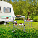 The DirectDeals Foldable Outdoor BBQ Table Grilling Stand is set with food, drinks, and utensils, with a white caravan on green grass in the background—your ideal camping cook station.