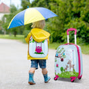 A child in a yellow raincoat stands beside the DirectDeals Fir Wood Kids Picnic Table with 2 Built-in Benches and Umbrella, along with a colorful animal suitcase, wearing rain boots on a wet outdoor path.