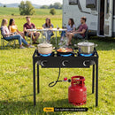 Five people relax by their RV in a grassy field while three pots cook on the DirectDeals 3 Burner Propane Stove 240000 BTU Outdoor Camping Grill with a red gas cylinder.