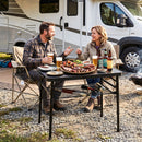 A man and woman enjoy grilled meats, beers, and sodas outdoors, smiling at a DirectDeals 3-Height Folding Camping Table with Large Storage Net & Handle near their RV.